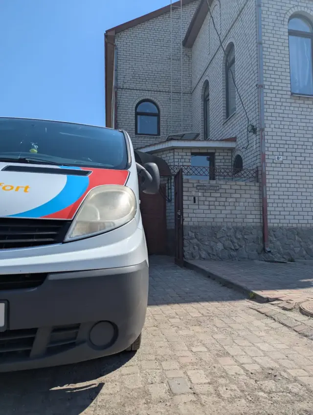 Передня частина білого мікроавтобуса медичного транспортування великим планом біля відчиненої хвіртки та воріт будинку з білої цегли з адресною табличкою.