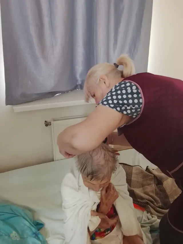 Доглядальниця в бордовому фартуху схилилася над літньою жінкою, загорнутою в білу тканину, і доглядає за її волоссям. Жінка сидить на ліжку біля вікна з сірою шторою та радіатором опалення.