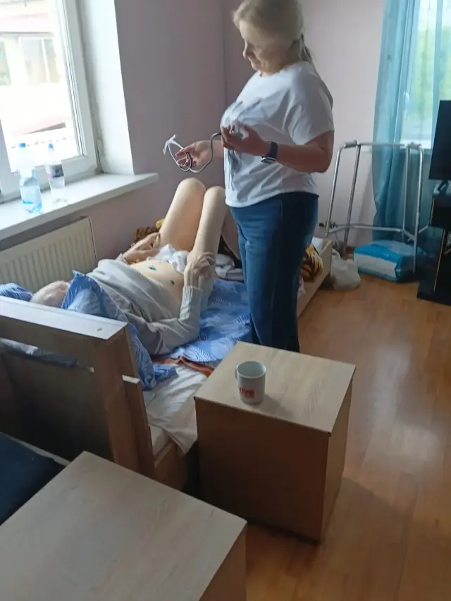 Доглядальниця у білій футболці стоїть із фонендоскопом біля ліжка літнього чоловіка, готуючись до огляду; світла кімната з великим вікном, радіатором опалення та ходунками для пересування біля стіни.
