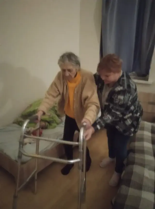 Доглядальниця підтримує літню жінку в бежевій кофті та жовтогарячому гольфі, допомагаючи їй підвестися з ліжка та стати на металеві ходунки. Дія відбувається у житловій кімнаті пансіонату.