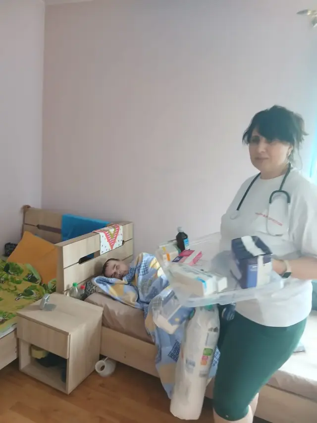Медична працівниця приносить лоток з медикаментами та пакунок гігієнічних засобів до кімнати, де на ліжку під блакитною ковдрою відпочиває чоловік. Світла кімната з дерев'яною тумбочкою.