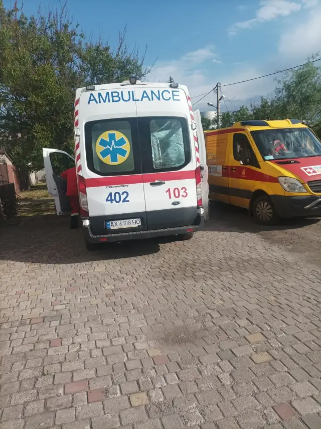 Дві карети швидкої допомоги на брукованому подвір'ї: білий автомобіль із написом AMBULANCE і номером 103 та жовто-червоний мікроавтобус приватної медичної служби; навколо дерева й літнє небо.