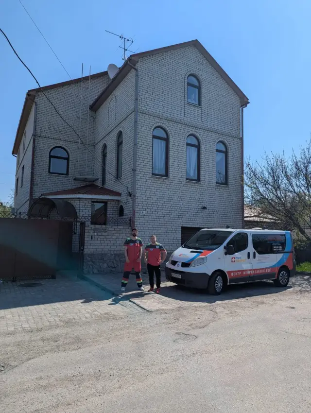 Двоє працівників служби перевезення хворих у червоній формі стоять поруч із білим брендованим мікроавтобусом перед триповерховою будівлею з білої цегли.