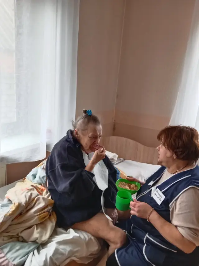 Доглядальниця у синьому фартусі сидить поруч із літньою жінкою на ліжку та подає їй обід — у руках зелена мисочка зі стравою та зелене горнятко. Жінка їсть, сидячи біля вікна з білими фіранками.