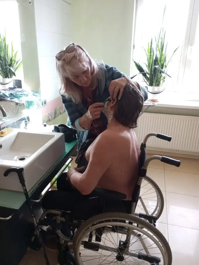 Доглядальниця акуратно голить літнього чоловіка, який сидить в інвалідному візку біля умивальника. Світла ванна кімната з дзеркалом, великим вікном і вазонами на підвіконні.