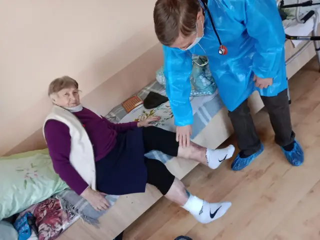 Лікар у блакитному захисному халаті та масці оглядає ногу літньої жінки, яка сидить на ліжку в кімнаті пансіонату. Поруч видно друге ліжко, ходунки для пересування та дерев'яну підлогу.