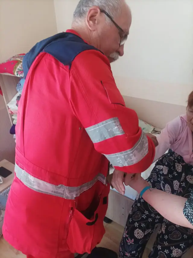 Лікар у червоній уніформі з світловідбивними смугами тримає за руку літню жінку в рожевій кофті, яка сидить на ліжку. Поруч тумбочка та полиці з речами в кімнаті пансіонату.