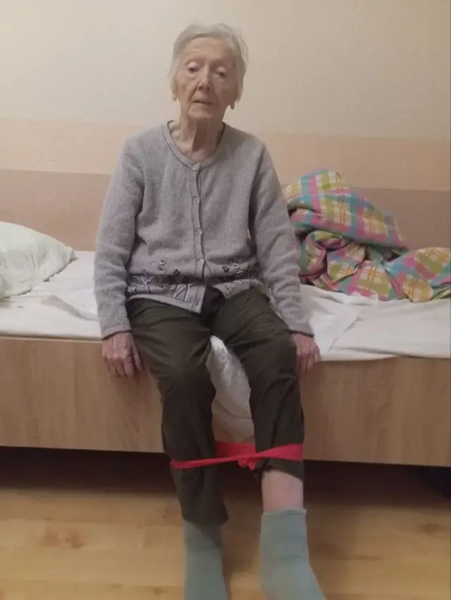 Пожилая женщина в серой кофте сидит на краю кровати с красной эластичной лентой на голенях во время реабилитационного упражнения для ног. Рядом на кровати сложенное цветное одеяло и подушка.