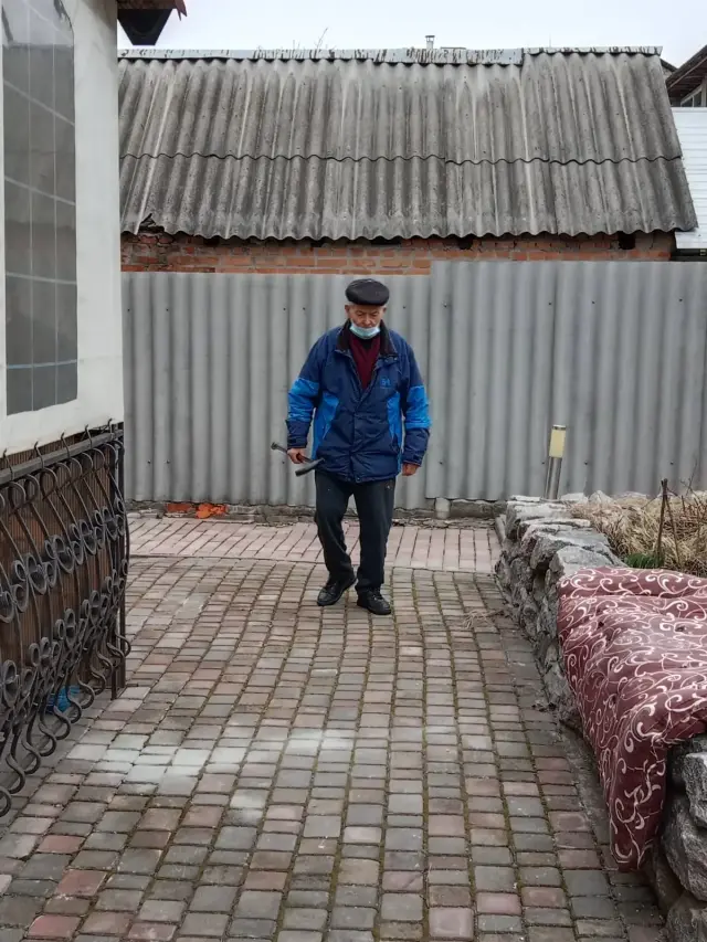 Пожилой мужчина в кепке и синей куртке прогуливается с палочкой по вымощенному брусчаткой двору. Вокруг ограда, каменная клумба и кованая решётка у здания.