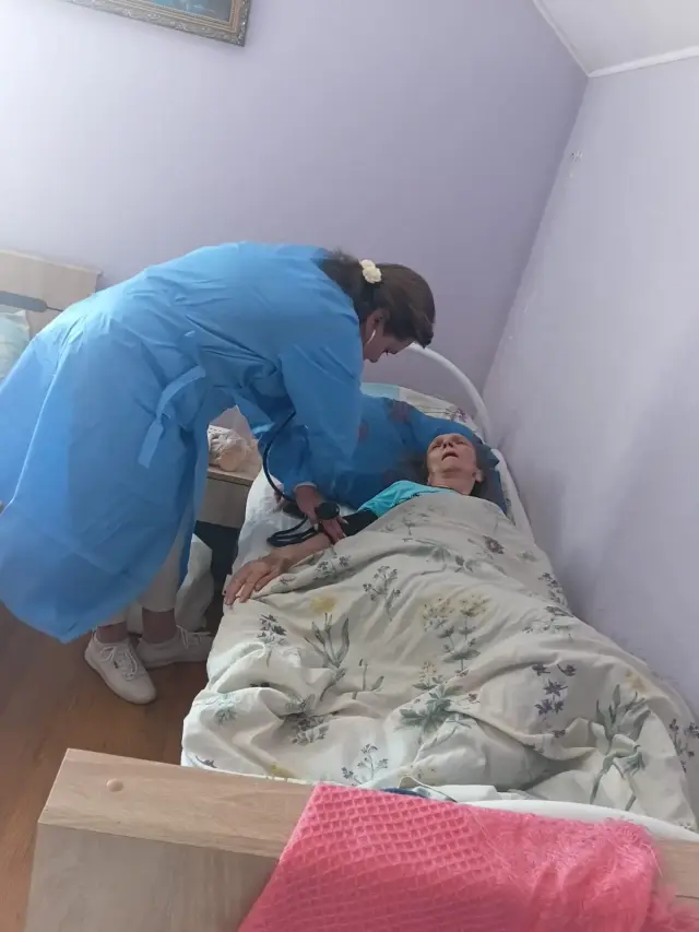 Доглядальниця у блакитному медичному халаті вимірює артеріальний тиск літній жінці, яка лежить у ліжку під ковдрою з квітковим візерунком; світла кімната з бузковими стінами та тумбочкою біля ліжка.