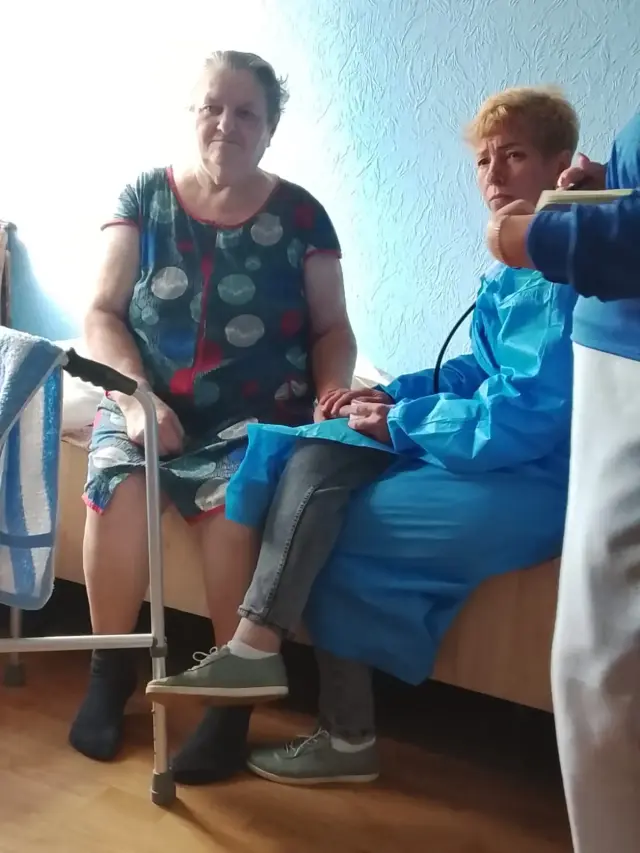 Літня жінка в сукні в горошок сидить на ліжку поруч із медпрацівницею в блакитному одноразовому халаті зі стетоскопом. Перед ліжком стоять ходунки для пересування, кімната зі світло-блакитними стінами.