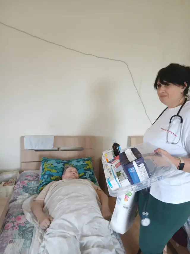 Медична працівниця зі стетоскопом тримає прозорий лоток з ліками та пакунок гігієнічних засобів біля ліжка, де під ковдрою лежить літня жінка з кольоровою подушкою під головою.