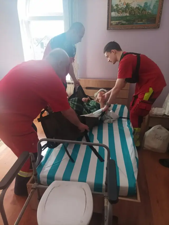 Троє медиків у червоній формі обережно перекладають літнього чоловіка з ліжка на м'які транспортувальні ноші у житловій кімнаті з картиною на стіні; на передньому плані крісло-туалет для маломобільних людей.