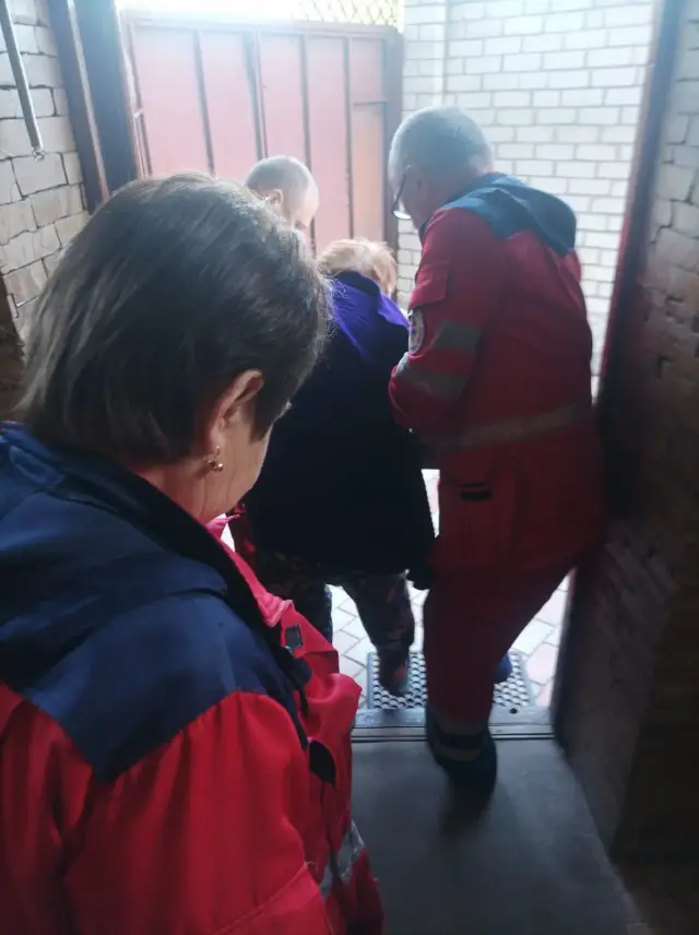 Двоє медиків у червоних уніформах швидкої допомоги обережно супроводжують літню жінку через вхідні двері цегляного будинку. Медики підтримують жінку з обох боків на сходах.