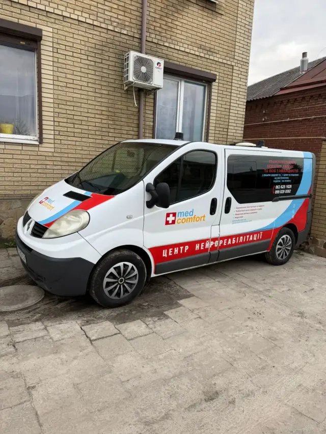 Білий мікроавтобус Renault Trafic з брендуванням med comfort і написом «Центр нейрореабілітації», призначений для транспортування лежачих хворих та осіб з інвалідністю, припаркований біля цегляної будівлі.