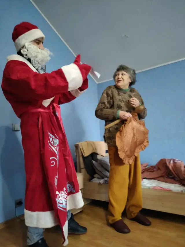 Аніматор у костюмі Діда Мороза вітає усміхнену літню жінку, яка стоїть посеред житлової кімнати пансіонату. Позаду видно ліжка з постіллю та рушниками на узголів'ї.