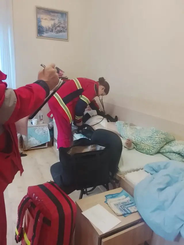 Фельдшерка в червоній уніформі зі стетоскопом схилилася над літньою людиною, що лежить у ліжку, а другий медик на передньому плані заповнює документи на планшеті. На підлозі стоїть червона медична валіза.