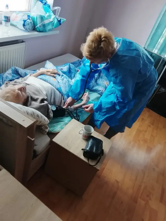 Медична працівниця у блакитному захисному халаті вимірює механічним тонометром тиск літній жінці, яка лежить у ліжку на блакитній постільній білизні; поруч тумбочка з чашкою та чохлом від тонометра, на підвіконні засоби гігієни.