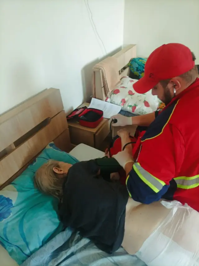 Парамедик у червоній формі та кепці вимірює артеріальний тиск літній людині, що лежить у ліжку на бірюзовій постільній білизні; на тумбочці — відкрита червона аптечка та аркуш із записами.