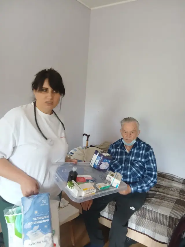 Медсестра зі стетоскопом тримає прозорий лоток із ліками та упаковку поглинаючих пелюшок перед літнім чоловіком у картатій сорочці. Чоловік сидить на ліжку з картатим покривалом у світлій кімнаті.