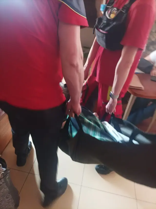 Двоє працівників екстреної медичної служби у червоній формі переносять пацієнта на м'яких ношах-волокушах коридором зі світлою плиткою на підлозі.