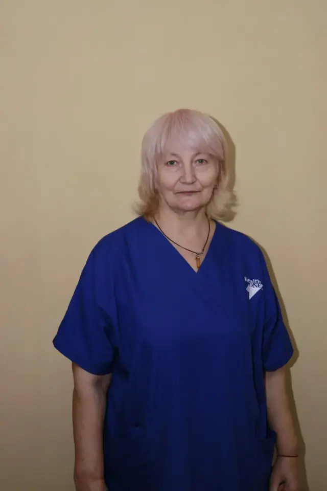 Женщина со светлыми волосами в тёмно-синем медицинском костюме с логотипом Health ONE стоит у бежевой стены. Портретное фото медицинской работницы пансионата.