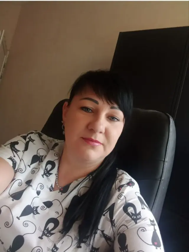 Женщина средних лет с тёмными волосами в светлой блузке с принтом котиков сидит в чёрном офисном кресле в кабинете. Портретное фото сотрудницы учреждения.