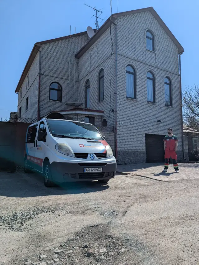 Мікроавтобус медичного транспортування стоїть перед триповерховим будинком з білої цегли, біля входу — працівник у червоній робочій формі. Сонячний весняний день.