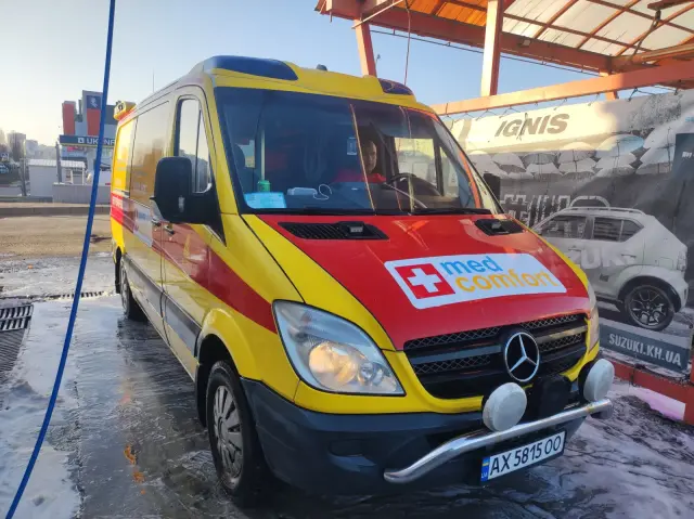 Жовто-червоний реанімобіль Mercedes-Benz Sprinter з логотипом med comfort стоїть на майданчику; у кабіні видно водія, на фоні — міська забудова.