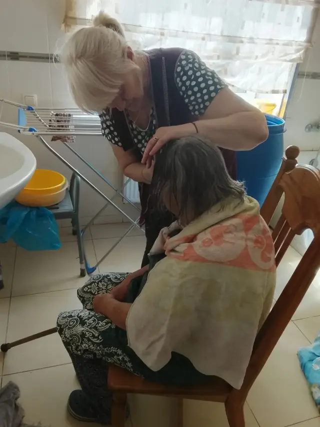 Доглядальниця розчісує вологе волосся літньої жінки, загорнутої в рушник, яка сидить на дерев'яному стільці у ванній кімнаті з плиткою. Поруч видно умивальник, жовтий таз, сушарку для білизни та душовий стільчик.
