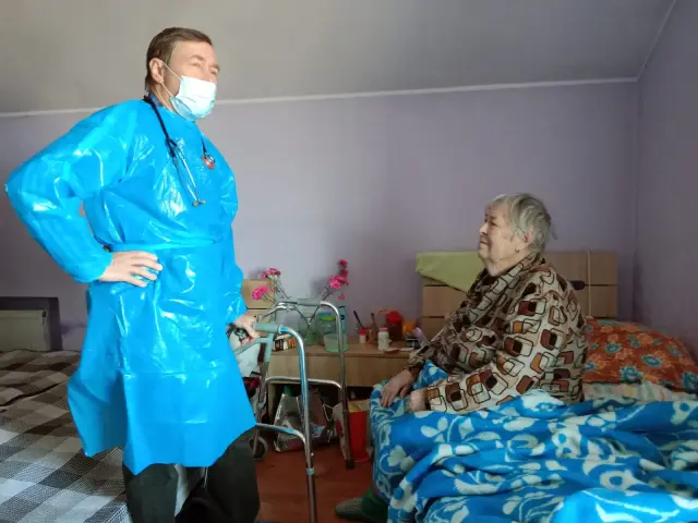 Лікар зі стетоскопом у блакитному халаті та масці спілкується з літньою жінкою, яка сидить у ліжку під ковдрою. У кімнаті видно ходунки, тумбочку з ліками та живі квіти.