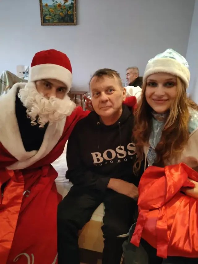 Мешканець пансіонату у чорному светрі сидить між аніматорами в костюмах Діда Мороза та Снігуроньки з червоним подарунковим мішком. Спільне святкове фото у житловій кімнаті, на задньому плані ще один літній чоловік.