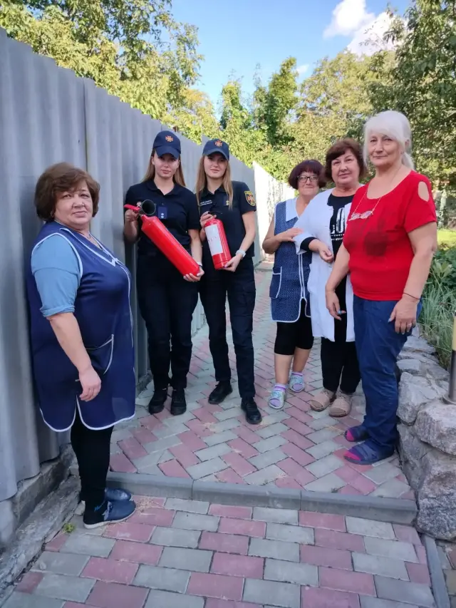 Дві співробітниці служби надзвичайних ситуацій у формі тримають червоні вогнегасники поруч із чотирма працівницями закладу на брукованій доріжці подвір'я. Навколо паркан, зелені дерева та сонячна погода.