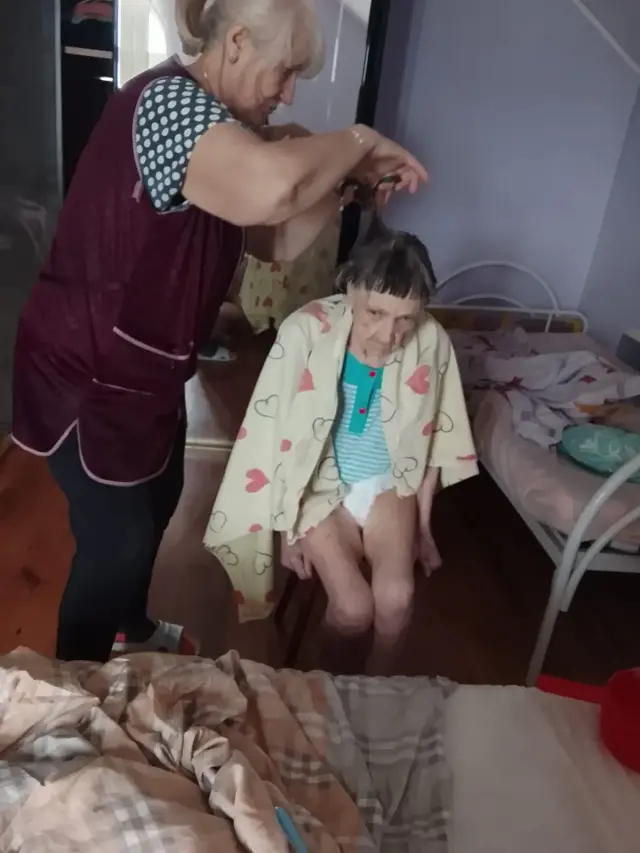 Доглядальниця в бордовому фартуху підстригає ножицями волосся літньої жінки, накритої світлою накидкою з сердечками. Жінка сидить на табуреті посеред кімнати, поруч ліжко з рожевою постільною білизною.