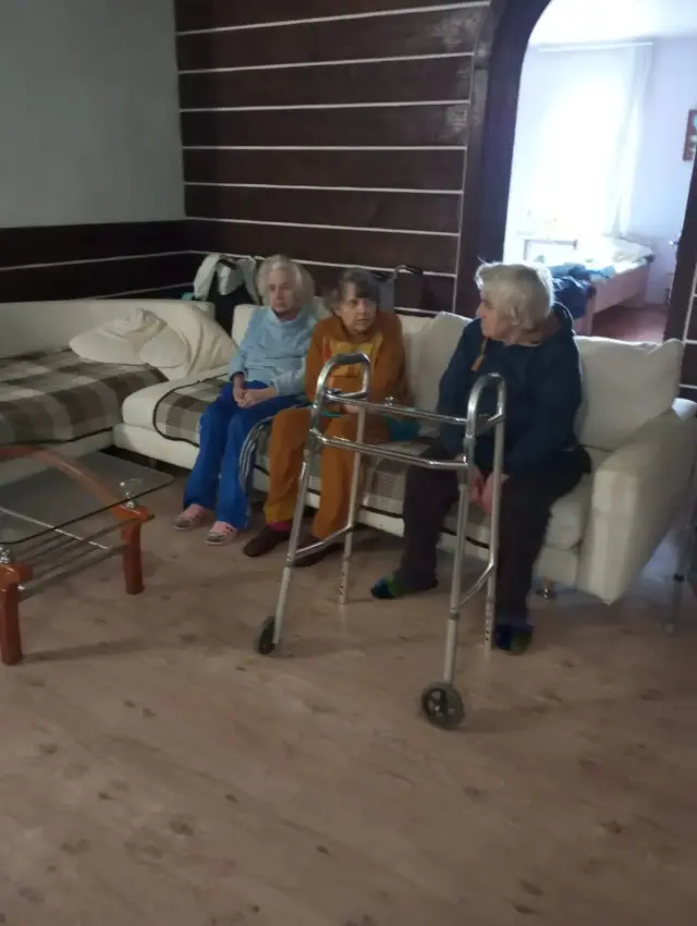 Троє літніх людей сидять на світлому дивані у вітальні з дерев'яним оздобленням стін, перед ними стоять ходунки на коліщатках. Поруч скляний журнальний столик, на задньому плані видно спальню.
