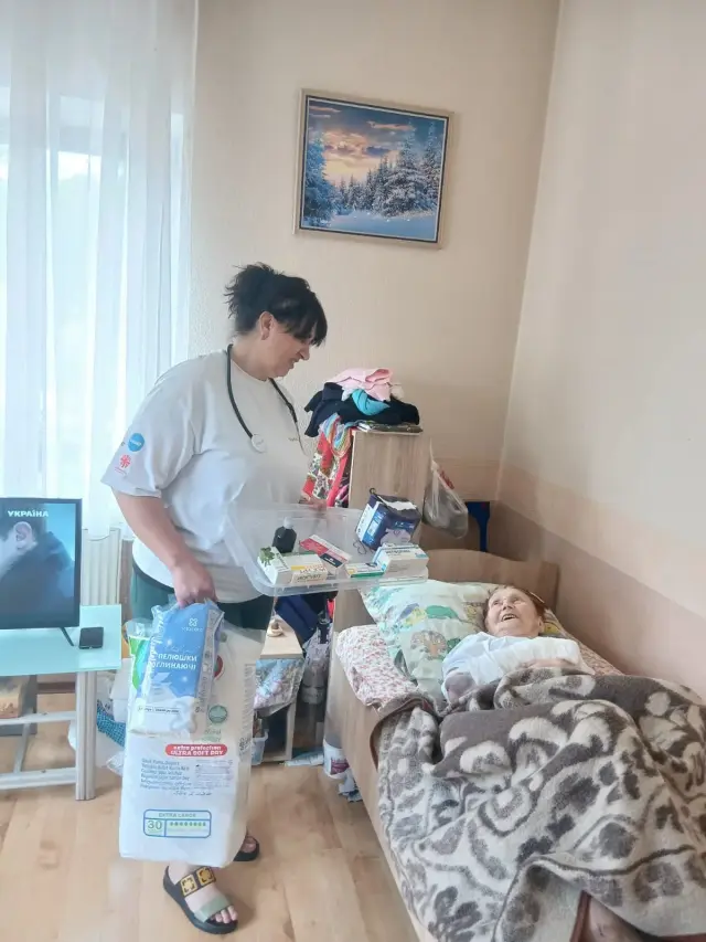 Медична працівниця з лотком медикаментів та упаковками поглинаючих пелюшок стоїть біля літньої жінки, яка лежить у ліжку та усміхається. У кімнаті телевізор і зимовий пейзаж на стіні.