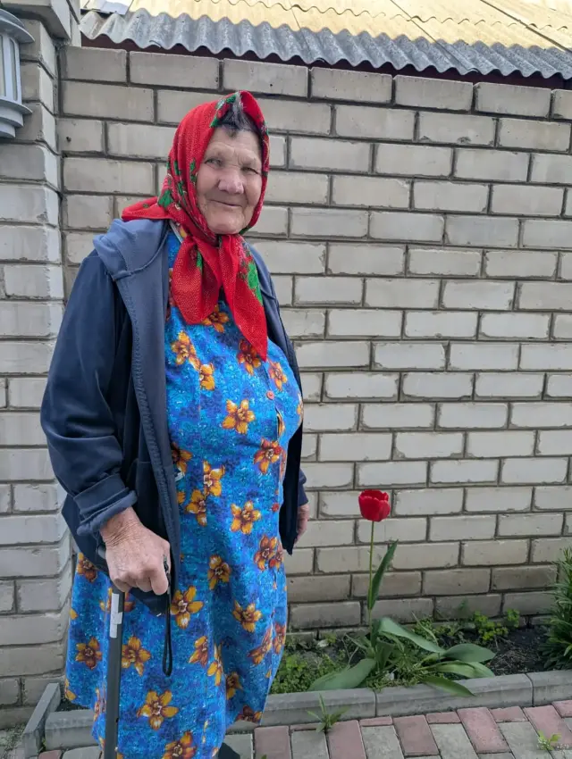 Усміхнена літня жінка в червоній хустці та синій квітчастій сукні стоїть з паличкою біля стіни з білої цегли. Поруч на клумбі квітне червоний тюльпан.