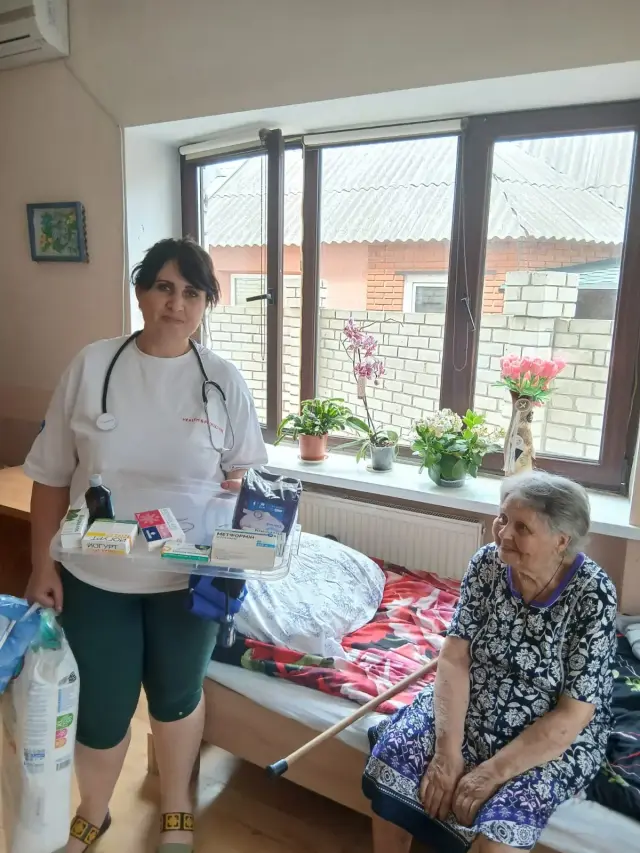 Медична працівниця тримає лоток з ліками та пакет засобів гігієни поруч із літньою жінкою, яка сидить на ліжку з паличкою та усміхається. На підвіконні — орхідея та квіти у вазонах.