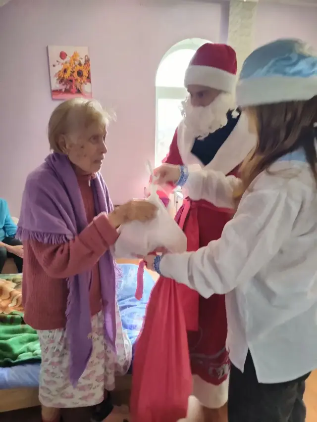 Дід Мороз і Снігуронька вручають літній жінці в бузковій шалі білий пакунок із подарунком у світлій кімнаті пансіонату. На стіні картина із соняшниками, поруч арочне вікно.