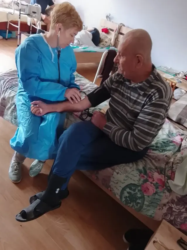 Медпрацівниця в блакитному захисному халаті вимірює артеріальний тиск літньому чоловікові в смугастій кофті, який сидить на ліжку з квітчастим покривалом. На задньому плані видно інші ліжка кімнати.