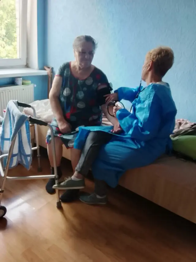 Медична працівниця у блакитному захисному халаті сидить на ліжку та вимірює артеріальний тиск літній жінці у сукні з квітковим принтом; на передньому плані ходунки з рушником, за вікном зелень.