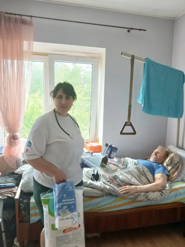 Медсестра в білій футболці тримає упаковку поглинаючих пелюшок біля ліжка усміхненого літнього чоловіка з надліжковою рамою-підйомником. На приліжковому столику розкладені ліки, за вікном видно зелені дерева.