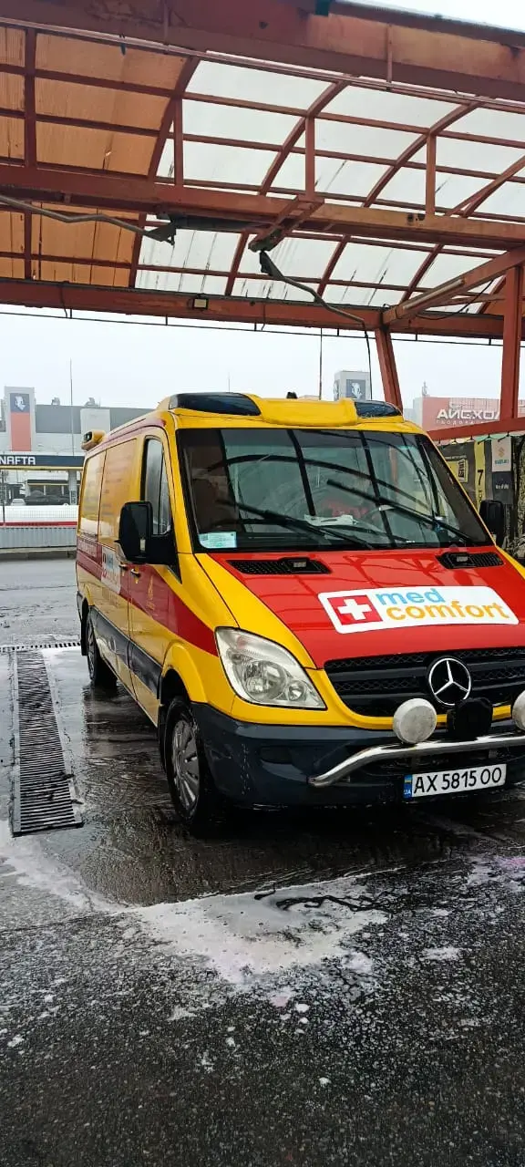 Жовто-червоний санітарний автомобіль Mercedes-Benz Sprinter з логотипом med comfort на капоті та додатковими фарами, припаркований під металевим навісом.
